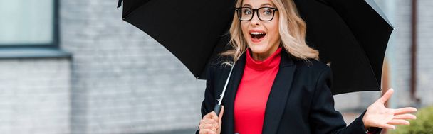 панорамный снимок привлекательной и потрясенной бизнесвумен в черном пальто с зонтиком в руках
  - Фото, изображение