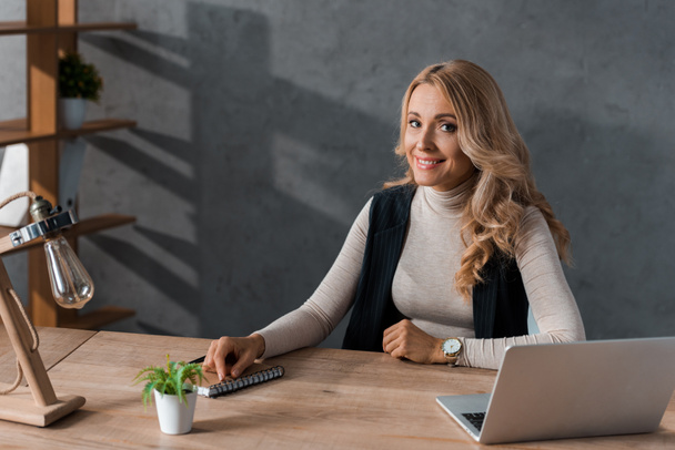 ελκυστική και ξανθιά επιχειρηματίας χαμογελά και κάθεται στο τραπέζι  - Φωτογραφία, εικόνα