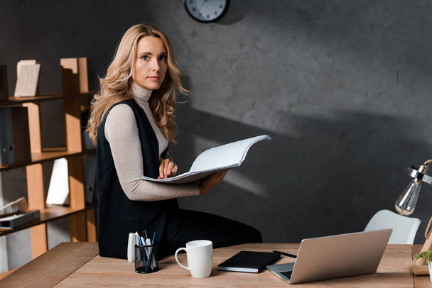attraktive und blonde Geschäftsfrau, die am Tisch sitzt und Papiere hält  - Foto, Bild