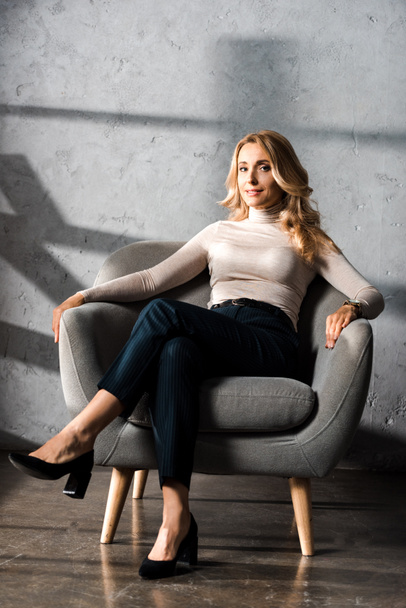 ελκυστική και ξανθιά επιχειρηματίας χαμογελά και κάθεται στην πολυθρόνα στο γραφείο  - Φωτογραφία, εικόνα