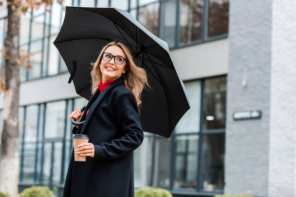 attractive and blonde woman in black coat holding paper cup and umbrella  - Foto, Imagem