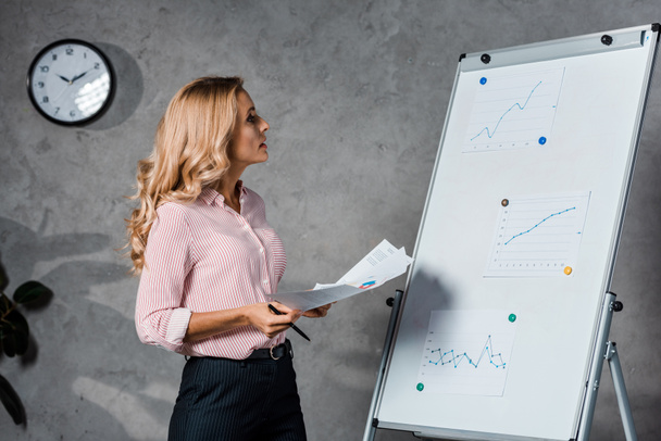 attractive and blonde businesswoman in shirt holding papers and looking at white board in office  - Foto, Bild