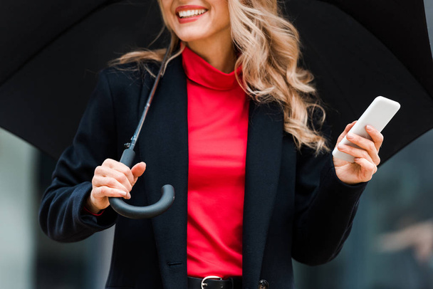 cropped view of blonde businesswoman in black coat smiling and holding smartphone  - Valokuva, kuva