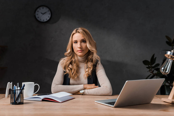 attractive and blonde businesswoman sitting at table and looking at camera in office  - Фото, зображення