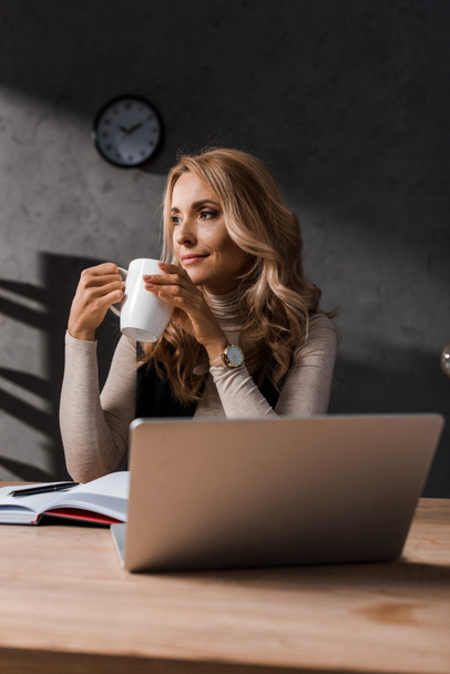 atraktivní a blond podnikatelka sedí u stolu a drží pohár  - Fotografie, Obrázek