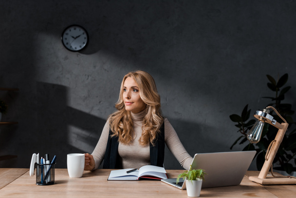 attractive and blonde businesswoman sitting at table and looking away in office  - 写真・画像