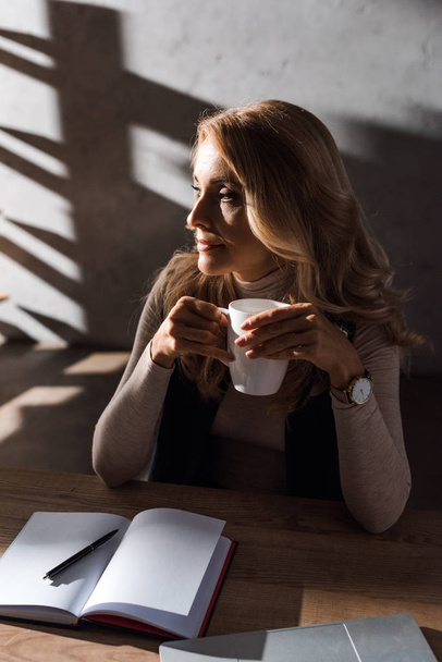 attraente e bionda donna d'affari in possesso di tazza e guardando lontano in ufficio
  - Foto, immagini