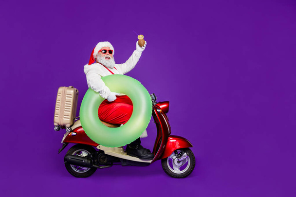 Full body photo of santa man going to meet newyear at tropics leaving cold december by bike hold life buoy and drink wear specs and costume isolated purple background - Фото, изображение