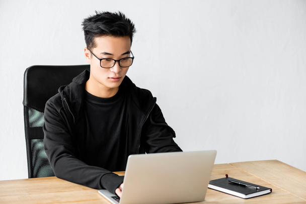 asian hacker in glasses sitting at table and using laptop  - Foto, Bild