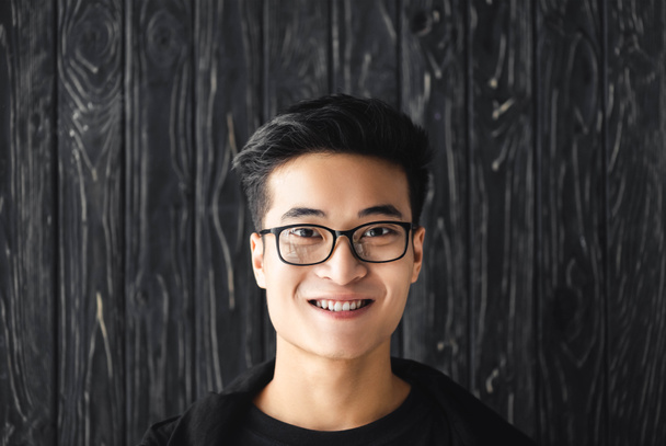 usmívající se asijský muž v brýlích při pohledu na kameru na dřevěném pozadí  - Fotografie, Obrázek