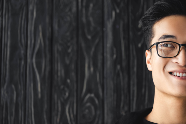 abgeschnittene Ansicht eines lächelnden asiatischen Mannes mit Brille, der auf hölzernem Hintergrund wegschaut  - Foto, Bild