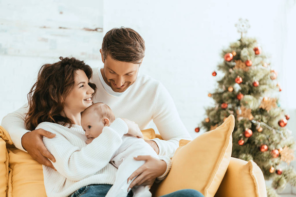 happy man hugging wife sitting on yellow sofa with baby near christmas tree - Photo, Image