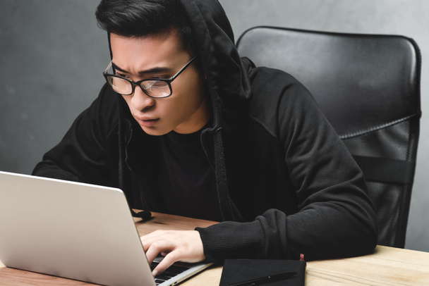 asian hacker in glasses using laptop and sitting at table  - Zdjęcie, obraz