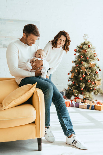 happy woman standing near christmas tree and looking at husband holding cute baby - Photo, Image