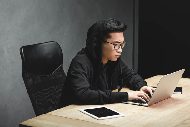 азіатський хакер в окулярах, використовуючи ноутбук і сидячи за столом
  - Фото, зображення