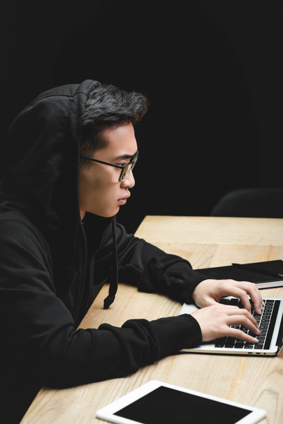 vista lateral de hacker asiático usando el ordenador portátil y sentado en la mesa
  - Foto, Imagen