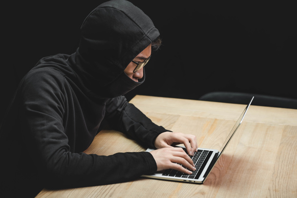 hoge hoek uitzicht van aziatische hacker met verduisterd gezicht met behulp van laptop en zitten aan tafel  - Foto, afbeelding