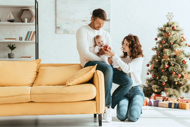 mujer atractiva dando bola de Navidad al bebé sentado en las manos de los padres cerca del árbol de Navidad decorado
 - Foto, Imagen