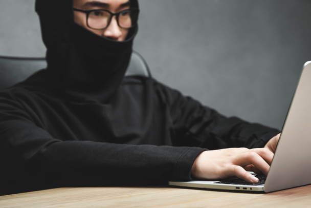 selective focus of asian hacker with obscured face using laptop and sitting at table  - Foto, afbeelding