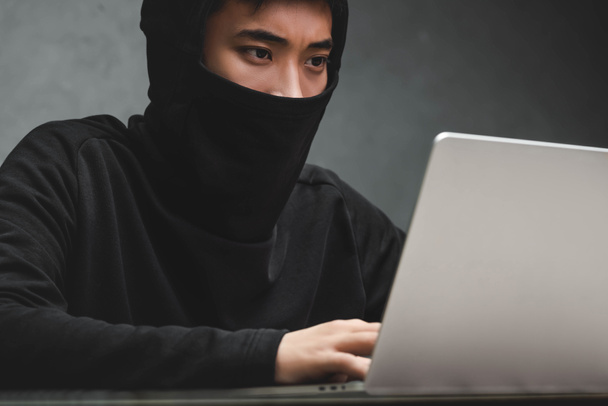 Suratı silik Asyalı hacker dizüstü bilgisayar kullanıyor ve masada oturuyor.  - Fotoğraf, Görsel