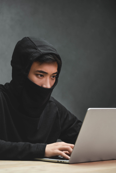 Ασιάτης χάκερ με καλυμμένο πρόσωπο που χρησιμοποιεί φορητό υπολογιστή και κάθεται στο τραπέζι  - Φωτογραφία, εικόνα
