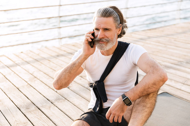 Image of pleased elderly man talking on cellphone while sitting on mat - Fotografie, Obrázek
