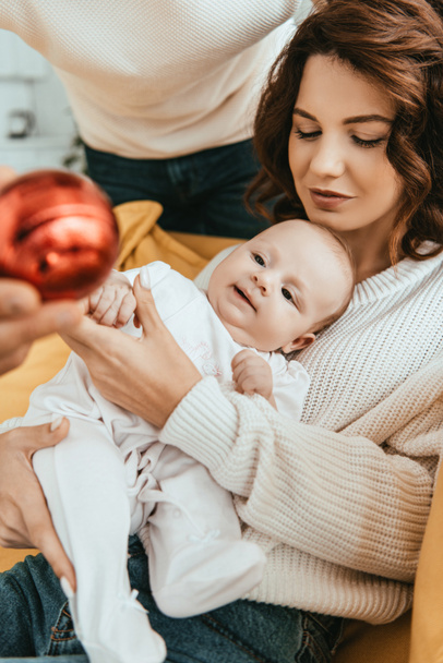 母親の手の上に横たわるかわいい幼児にクリスマスボールを示す父親のクロップドビュー - 写真・画像