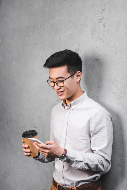 sonriente asiático hombre de negocios celebración de papel taza y el uso de smartphone
  - Foto, Imagen