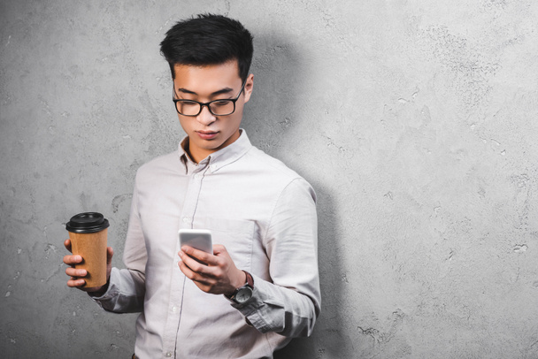 asian businessman holding paper cup and using smartphone in office  - Foto, Bild