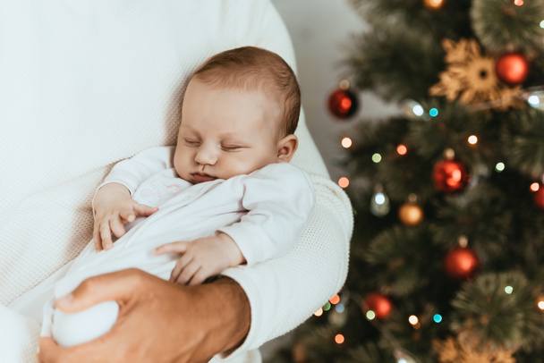обрезанный вид отца, держащего спящего ребенка возле рождественской елки
 - Фото, изображение