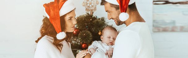 happy woman looking at smiling husband holding little daughter near christmas tree - Photo, Image