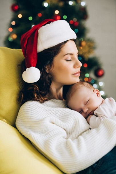 young woman in santa hat sitting with closed eyes and holding sleeping baby - Fotoğraf, Görsel