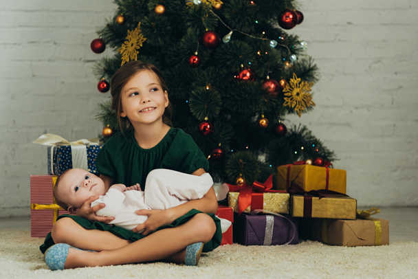 счастливый ребенок, держащий милого младенца, сидя рядом с елкой и коробками с подарками
 - Фото, изображение