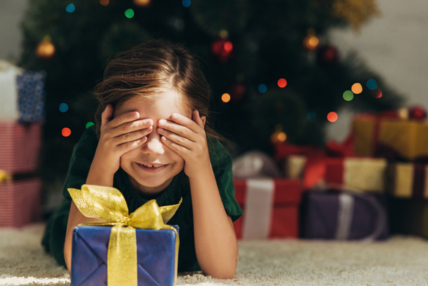 smiling kid lying on floor near gift box and covering eyes with hands - 写真・画像
