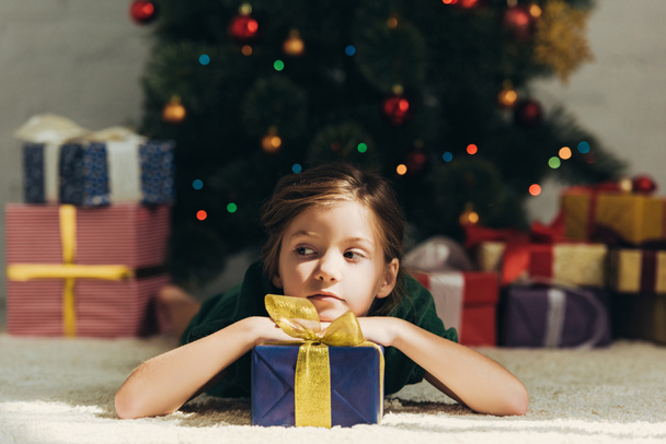 dreamy child looking away while lying on floor near gift box and christmas tree - Φωτογραφία, εικόνα