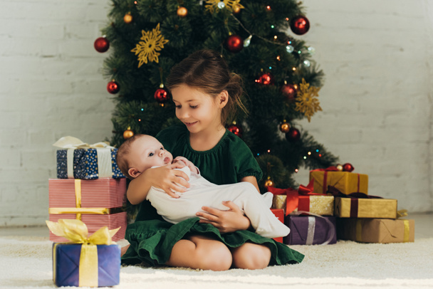 αξιολάτρευτο παιδί κρατώντας χαριτωμένο μωρό, ενώ κάθεται κοντά στο χριστουγεννιάτικο δέντρο και κουτιά δώρων - Φωτογραφία, εικόνα