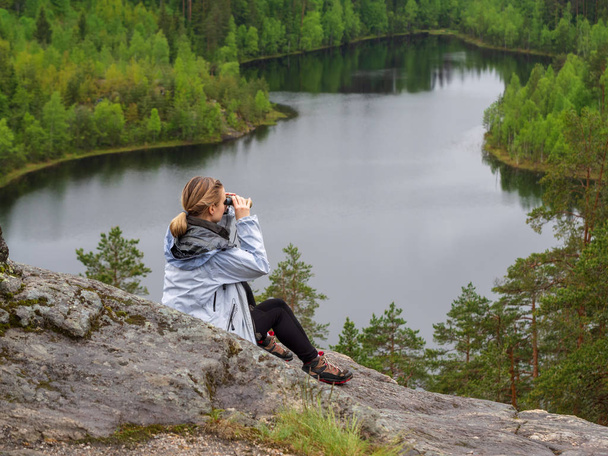 woman looks through binoculars. tourist girl on a rock in a forest near a lake - Photo, image