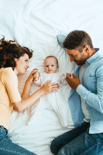 happy parents gently touching adorable baby while lying on white bedding together - Photo, Image