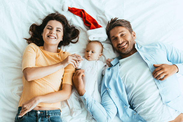 top view of happy parents lying on bedding near baby and santa hat - Foto, Imagem