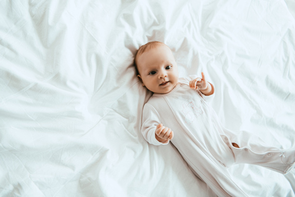 top view of cute infant in white bodysuit lying on bedding at home - Zdjęcie, obraz