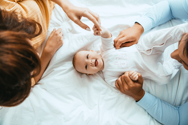top view of parents touching smiling infant lying on white bedding - Fotoğraf, Görsel