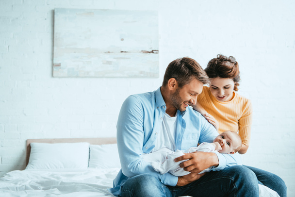 happy man holding little baby while sitting on bed near smiling wife - Foto, Imagem