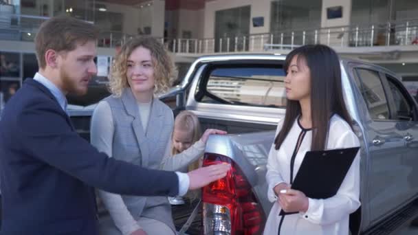 family auto buying, car dealer worker helps to choose vehicle for customers young couple with child in trunk of new machine at car sales center - Footage, Video