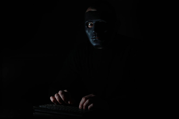 ein Mann mit Maske drückt im Dunkeln auf die Tastatur - Foto, Bild