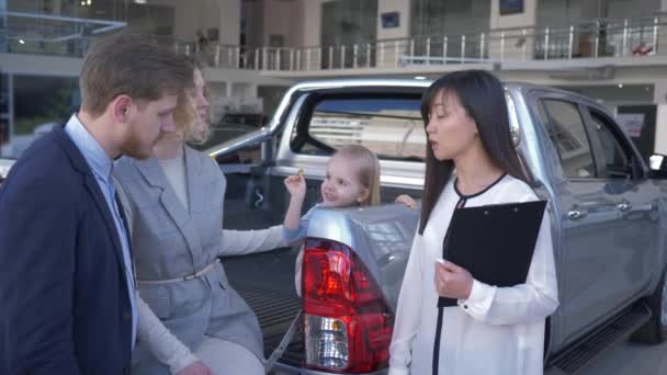 щаслива покупка автомобіля, продавець автомобілів консультуватися з покупцями щаслива сім'я з маленькою донькою в багажнику автомобіля в центрі продажу
 - Кадри, відео