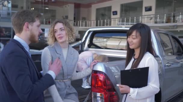 szczęśliwy sprzedawca samochodów doradza kupującym rodzina z dzieckiem dziewczyna w bagażniku pojazdu i uścisnąć dłonie po udanej sprzedaży w Auto centrum sprzedaży - Materiał filmowy, wideo