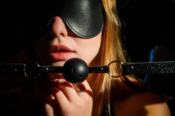 A man dominates and puts a gag in her mouth. BDSM concept. Portrait of a woman in seductive underwear with an intimate toy in her mouth. Sexy couple plays love games. Eyes closed with a leather mask. - Photo, Image