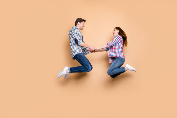 Full length profile photo of two people crazy lady guy jumping high holding hands cheerful playful mood wear casual plaid jeans clothes isolated beige color background - Foto, afbeelding