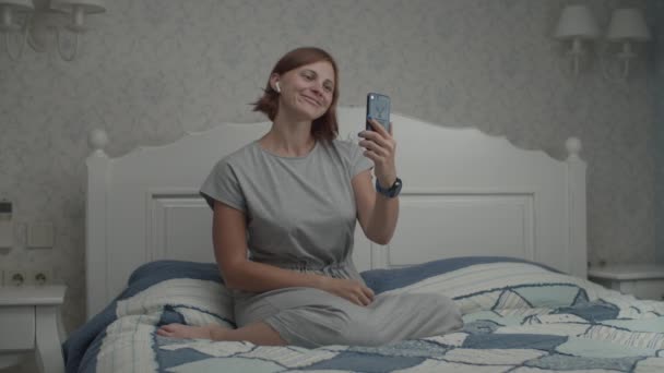 Slow motion of young pretty women in grey dress and wireless earphones admires her reflection in smartphone camera. Female taking selfie on phone in bedroom - Imágenes, Vídeo