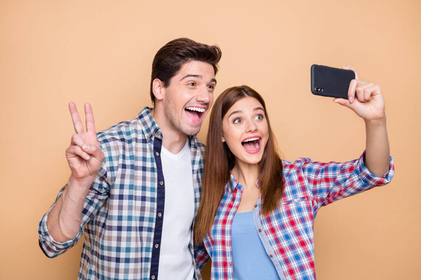 Close-up portrait of his he her she nice attractive lovely funky cheerful cheery couple wearing checked shirt taking selfie showing v-sign isolated over beige pastel color background - Fotó, kép